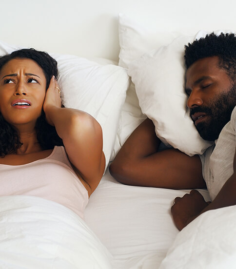 woman covering her ears as her sleeping husband snores next to her